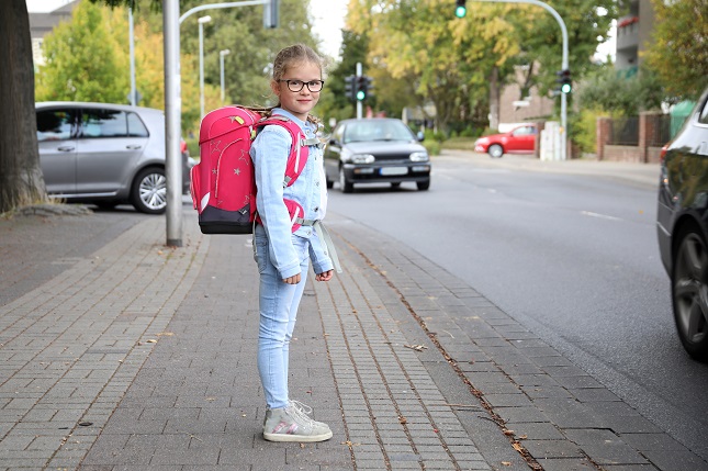 Mädchen mit Schulranzen an einer Straße im Hintergrund ein Auto